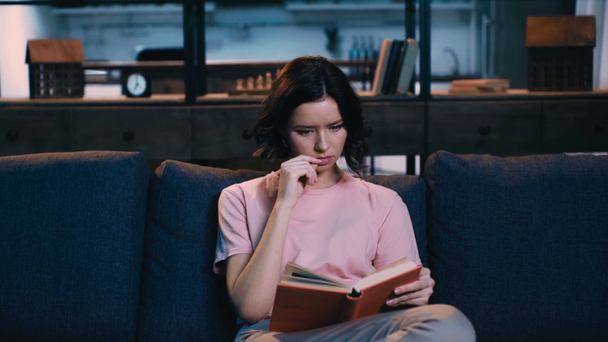 szép, figyelmes nő ül a kanapén otthon és olvasott könyv - Felvétel, videó