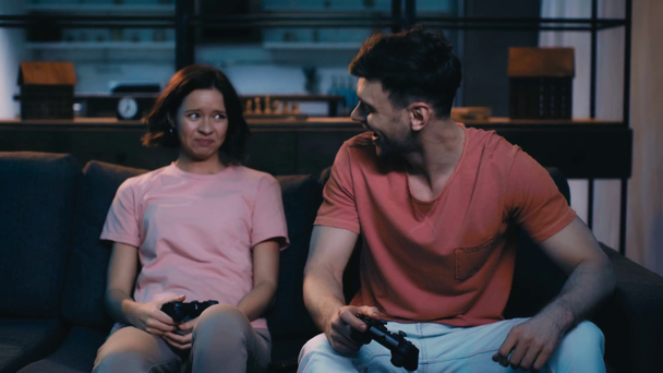 KYIV, UKRAINE - APRIL 12, 2019: Excited man and woman playing video game with joysticks while sitting on sofa at home. - Materiaali, video