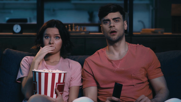 wstrząśnięty młoda kobieta z papieru kubek popcorn i młody człowiek z pilotem do oglądania telewizji, siedząc na kanapie w domu - Materiał filmowy, wideo