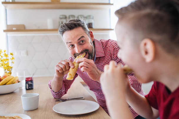 Mature father with small son indoors sitting at the table, eating pancakes. - Photo, Image