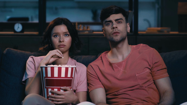 wstrząśnięty młody mężczyzna i kobieta oglądanie telewizji, jedzenie popcorn i patrząc na siebie, siedząc na kanapie w domu - Materiał filmowy, wideo