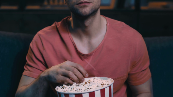 Teilbild eines jungen Mannes, der Popcorn isst, während er zu Hause auf der Couch sitzt und fernsieht - Filmmaterial, Video