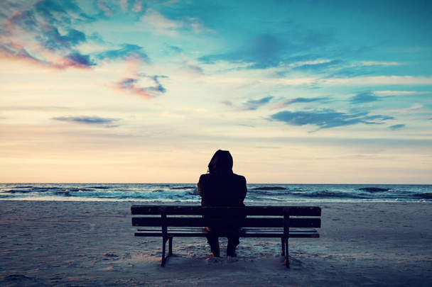 Férfi sziluettje kapucnival ül a padon a tengerparton naplementekor - Fotó, kép