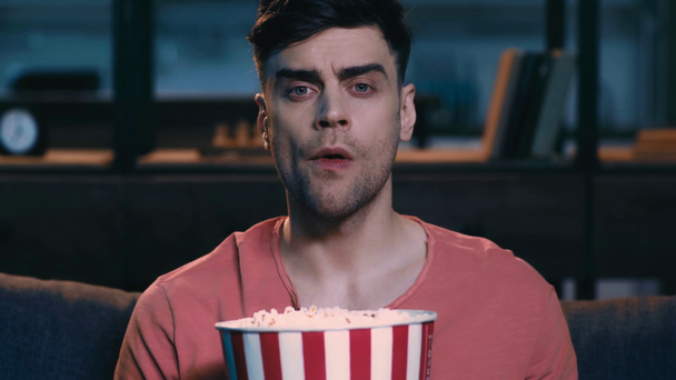 verrast jonge man kijken tv terwijl zittend op de Bank thuis en het houden van papieren beker met popcorn - Video