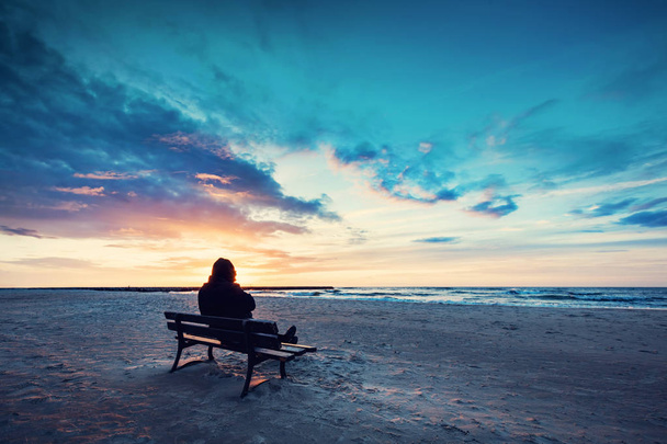 Męska sylwetka w kaptur siedzi na ławce na plaży o zachodzie słońca - Zdjęcie, obraz