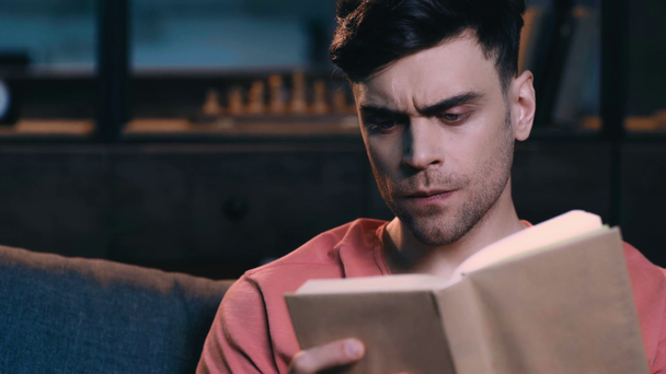 serious young man sitting on sofa at home and reading book - Imágenes, Vídeo