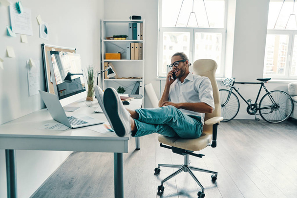 businessman in shirt talking on the smart phone and smiling while sitting in the office with legs on desk  - Photo, Image