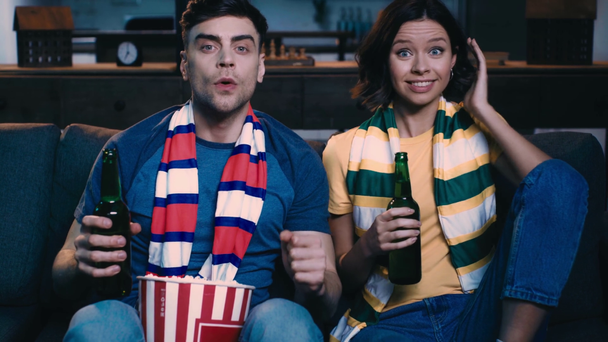 自宅のソファで動揺した男の近くでサッカーの試合を見ながら勝者のジェスチャーを示す興奮した若い女性 - 映像、動画