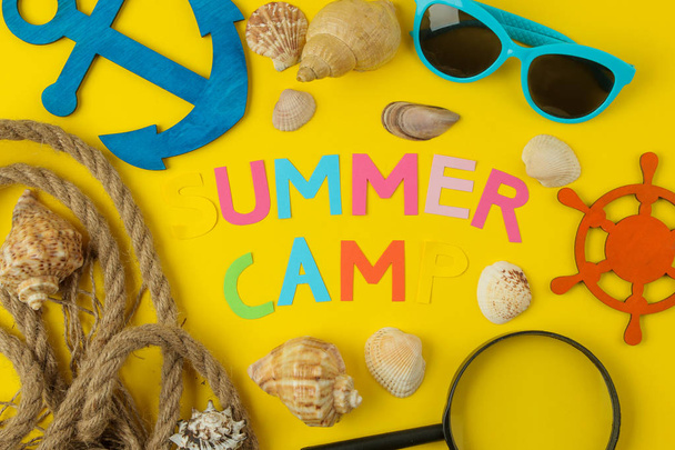 Tekst zomerkamp van veelkleurige papieren brieven en schelpen en een decoratief stuurwiel op een heldere gele achtergrond. bovenaanzicht. platte lay - Foto, afbeelding