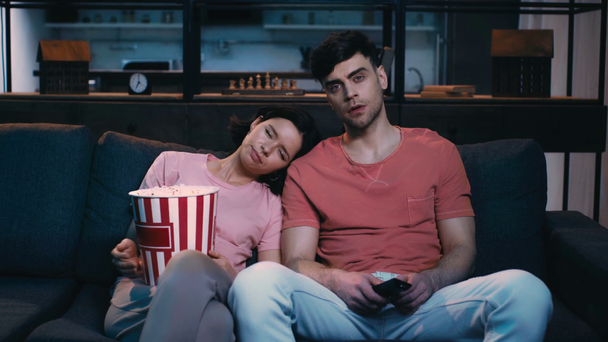 young woman holding paper cup with popcorn and yawning while leaning of shoulder of man holding remote controller and watching tv at home - Footage, Video