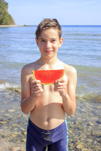Χαριτωμένο έφηβο αγόρι κρατά ένα κομμάτι καρπούζι στην παραλία δίπλα στη θάλασσα - Φωτογραφία, εικόνα