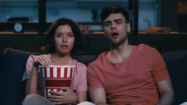 überrascht Mädchen Popcorn essen, während auf der Couch sitzen und Fernsehen in der Nähe schockierten jungen Mann - Filmmaterial, Video