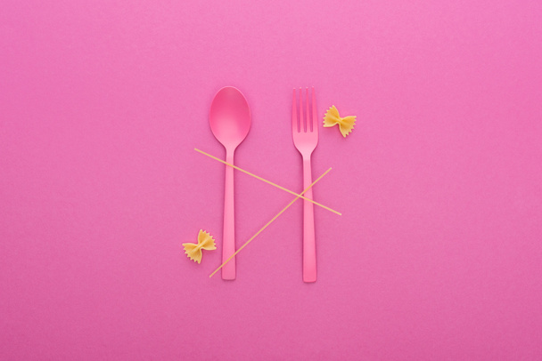 pink plastic spoon and fork, spaghetti and two uncooked farfalle pasta isolated on pink - 写真・画像
