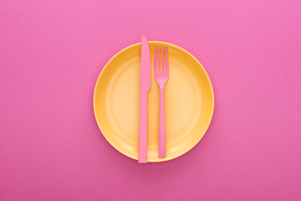 top view of pink plastic fork and knife on yellow plastic plate on pink background - Fotoğraf, Görsel