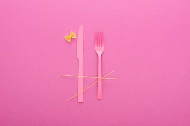 růžový plastikový nůž a vidlička, špagety a tepelně neupravené těstoviny izolované na růžovém - Fotografie, Obrázek