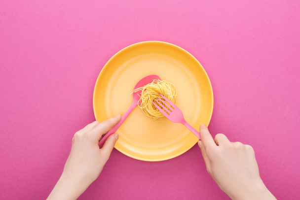 cropped view of adult holding pink spoon and taking vermicelli pasta with fork on yellow plastic plate on pink background - Photo, Image