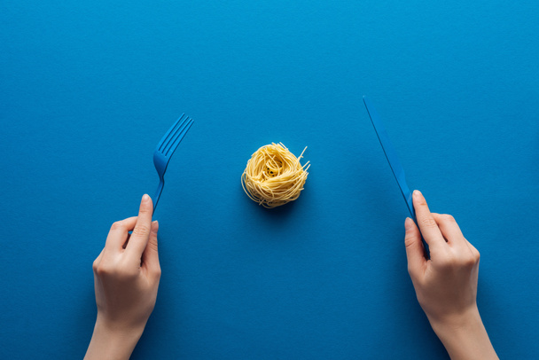 cropped view of woman holding blue plastic fork and knife with vermicelli pasta in middle on blue background - Photo, Image