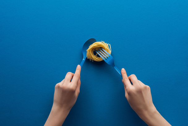 cropped view of woman holding blue spoon and taking vermicelli pasta with fork isolated on blue - Valokuva, kuva