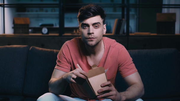 attente jonge man kijken tv terwijl zittend op de Bank thuis en het eten van een hapje met houten stokken - Video