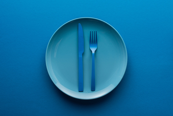 blue plastic knife and fork on plate on blue background - 写真・画像
