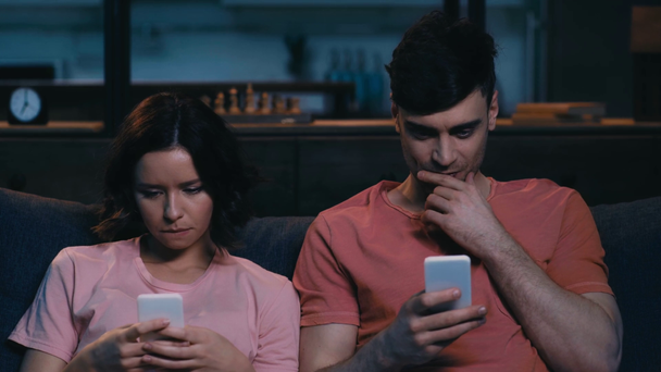 alegre joven hombre y mujer usando teléfonos inteligentes, hablando y sonriendo mientras está sentado en el sofá en casa
 - Metraje, vídeo