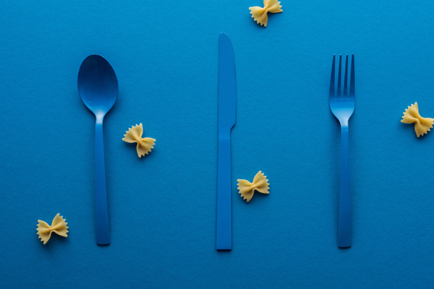 blue plastic spoon, knife and fork with uncooked farfalle pasta around on blue background - Photo, Image