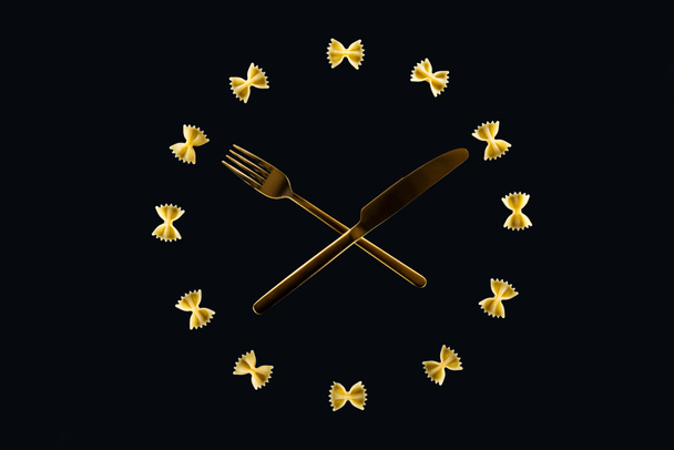 pohled na hodinový koncept s těstovinovými těstoviny, zkříženým vidličkou a nožem uprostřed izolované na černém - Fotografie, Obrázek