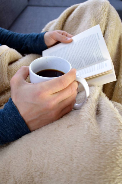 Um jovem está sentado em um sofá e lendo um livro de close-up sobre o livro e copo de vidro cheio de chá conceito para aquecer
  - Foto, Imagem