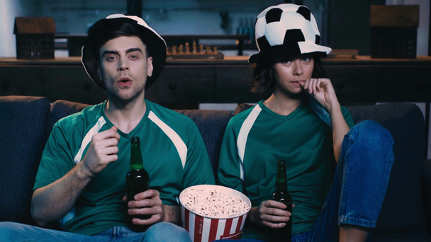 ενθουσιασμένος άντρας και γυναίκα σε καπέλα ανεμιστήρα πίνοντας μπύρα και τρώγοντας ποπ κορν, παρακολουθώντας ποδόσφαιρο αγώνα στην τηλεόραση και δείχνοντας χειρονομίες θριάμβου - Πλάνα, βίντεο