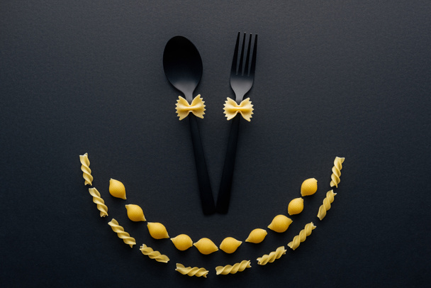 uncooked farfalle pasta on plastic spoon and fork near shells macaroni and rotini pasta isolated on black - Fotó, kép