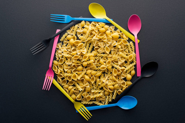 top view of uncooked pasta surrounded by colorful spoons and forks on black background - Photo, Image