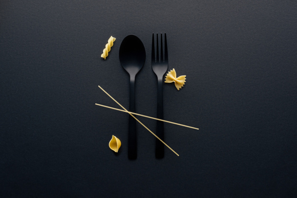 black plastic spoon, fork and four different kinds of pasta on black background - Fotó, kép