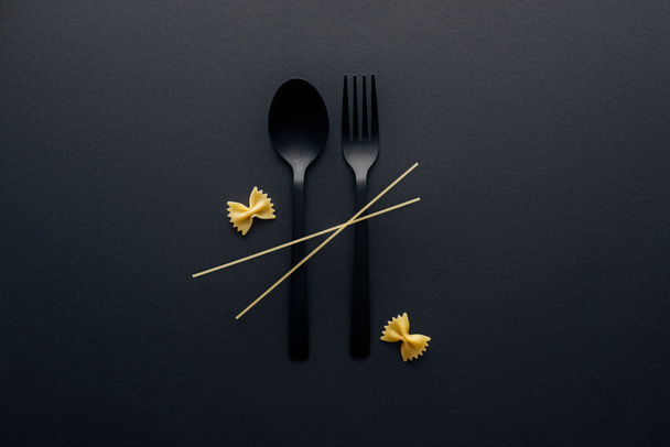 plastic lepel en vork met Farfalle pasta en spaghetti op zwarte achtergrond - Foto, afbeelding