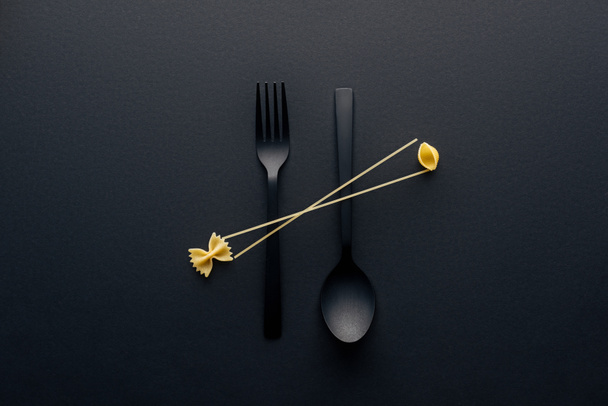 flat lay of spaghetti, farfalle pasta and shell macaroni on spoon upside down and fork on black background - Foto, Imagem