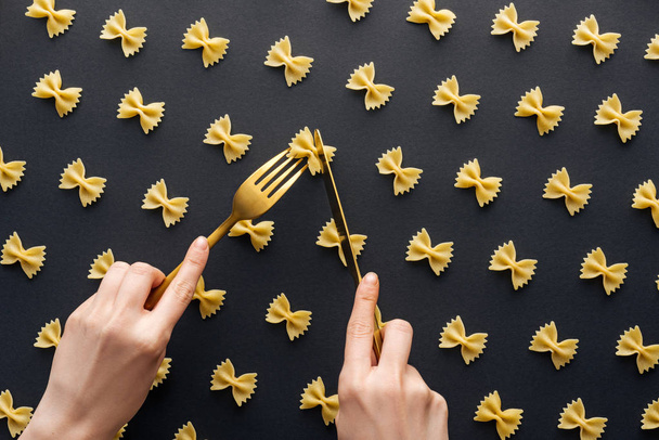 cropped view of woman holding yellow fork and cutting farfalle pasta with knife isolated on black - Фото, зображення