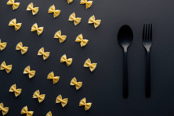 flat lay of farfalle pasta near plastic spoon and fork on black background - Valokuva, kuva