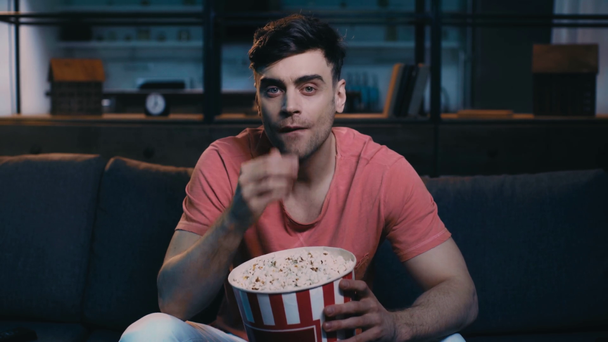 attente jonge man die popcorn eet, glimlacht en verraste emotie toont terwijl hij thuis tv kijkt - Video