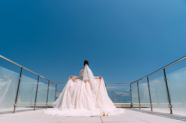 Backside view of bride stands on balcony in white dress with long train - Photo, image