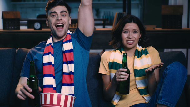 自宅でテレビでサッカーの試合を見て、ビールを飲み、ポップコーンを食べて、男性が動揺した女の子の近くに勝者のジェスチャーを示すとき、ストライプのスカーフの若いファン - 映像、動画