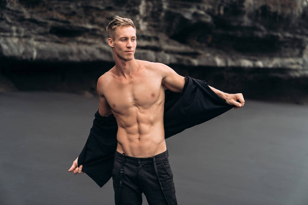 Sexy fitness male model in black pants and shirtless posing on black sandy beach. - Fotografie, Obrázek