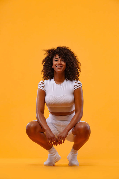 Smiling strong athletic woman with black skin and curly hair, doing exercise on yellow background wearing sportswear. Fitness and sport motivation. - Foto, afbeelding