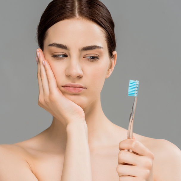 attractive naked woman touching face and looking at toothbrush isolated on grey  - Foto, imagen