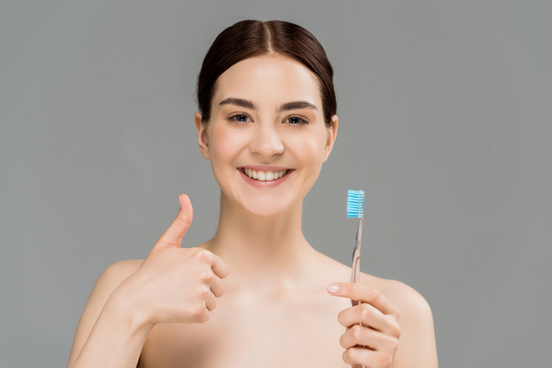 cheerful naked woman holding toothbrush while showing thumb up isolated on grey  - 写真・画像