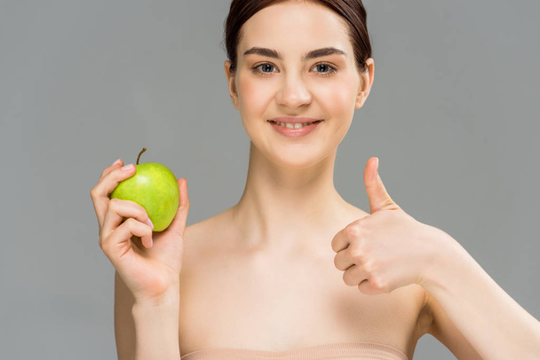 happy woman smiling while holding green apple and showing thumb up isolated on grey  - 写真・画像