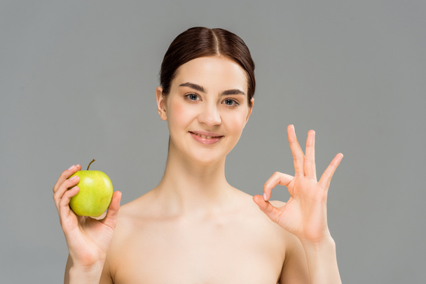 alegre mujer desnuda sonriendo mientras sostiene la manzana verde y mostrando signo ok aislado en gris
  - Foto, Imagen