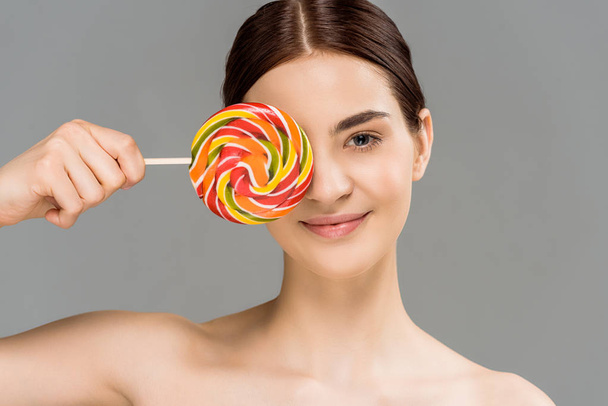 vrolijke naakte vrouw bedekt oog met kleurrijke Lollipop geïsoleerd op grijs  - Foto, afbeelding