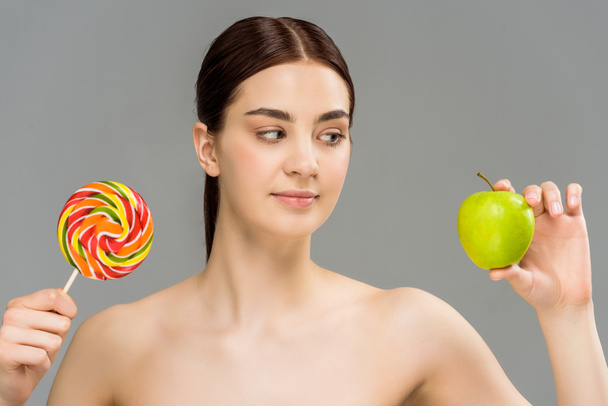 aantrekkelijk naakt meisje kijkend naar groene appel terwijl het houden van zoete Lollipop geïsoleerd op grijs  - Foto, afbeelding