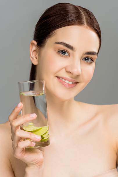 Veselá dívka držící sklenici vody s nakrájeným vápnem izolovaným na šedé  - Fotografie, Obrázek