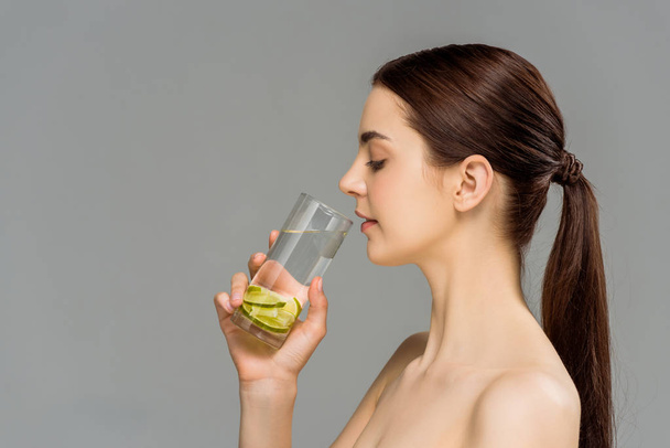 vista laterale di attraente donna nuda guardando vetro di acqua con calce tagliata isolata su grigio
  - Foto, immagini