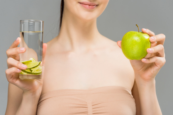 bijgesneden weergave van vrolijke meisje met glas water met gesneden kalk en groene appel geïsoleerd op grijs  - Foto, afbeelding